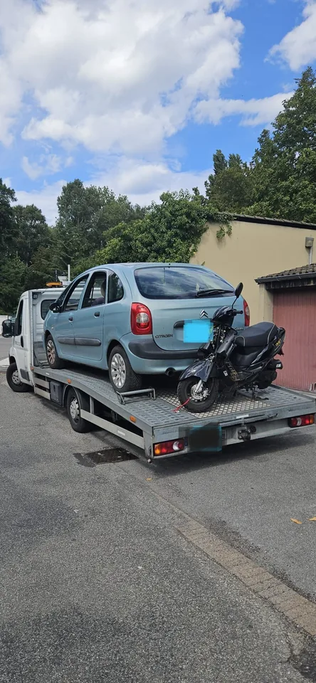 Citroën Xsara Picasso et scooter sur le plateau d’un camion dans une rue.