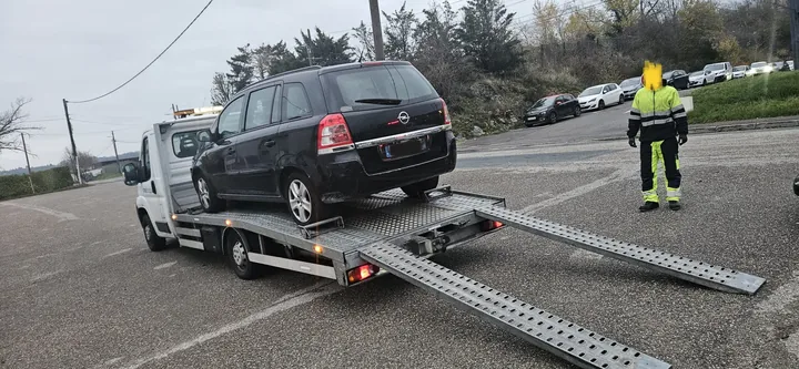 Véhicule familial sombre sur un camion plateau avec deux rampes abaissées et une personne à proximité.