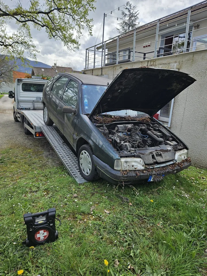 Voiture sombre au capot ouvert sur l’herbe devant un camion plateau.