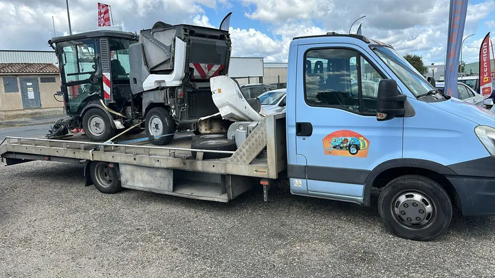 Balayeuse compacte avec cabine et brosses chargée sur un camion plateau blanc.