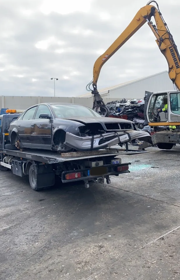 Berline incomplète sur un plateau, sans plusieurs roues ni éléments avant, près d’un engin à pince.